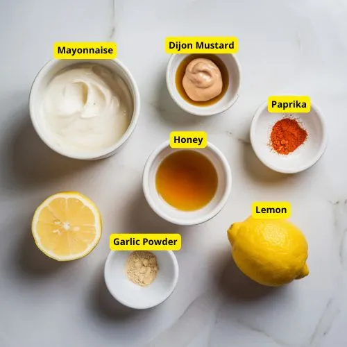 Ingredients for homemade chicken sauce on a white marble surface, including mayonnaise, Dijon mustard, honey, lemon juice, paprika, garlic powder, and a kitchen whisk.